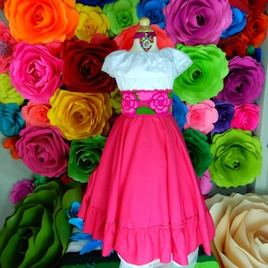 May include: A pink skirt with a green and pink embroidered belt, a white blouse with lace trim, and a pink flower necklace. The skirt has a ruffled hem and is worn on a mannequin against a background of colorful paper flowers.