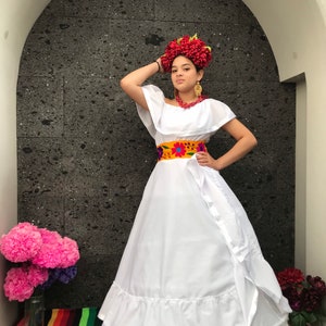 Pode incluir: Um vestido branco com saia rodada e cinto bordado. O vestido é usado por uma pessoa com uma coroa de flores vermelhas e brincos. A pessoa está em pé em frente a uma parede de pedra cinza.