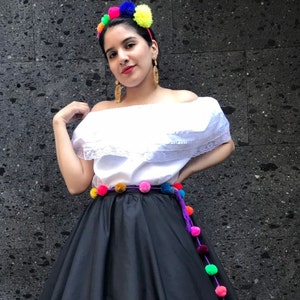 May include: A woman wearing a traditional Mexican outfit. The outfit includes a white off-the-shoulder blouse with lace trim, a black skirt, and a colorful pom-pom belt. A headband with colorful pom-poms and gold earrings complete the look.
