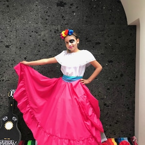 May include: A young girl dressed in a traditional Mexican costume for Dia de los Muertos. She is wearing a white blouse with a ruffled collar, a pink skirt with ruffles, and a blue sash. She has a flower crown on her head and a sugar skull painted on her face.