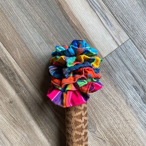 May include: A wooden rolling pin with a carved pattern, topped with a colorful fabric ruffle. The fabric is a mix of bright colors, including pink, orange, yellow, blue, and green.