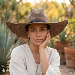 May include: A brown woven straw cowboy hat with a wide brim and a dark brown leather band featuring a central circular metal accent. The hat has a structured crown with a pinched top and a textured finish, providing a classic western appearance.