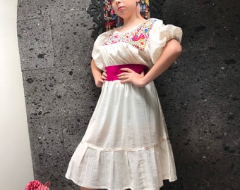 May include: A white cotton dress with embroidered details and a pink sash. The dress has a ruffled hem and puffed sleeves. The model is wearing a pink headband and sandals.