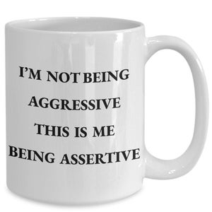May include: White ceramic coffee mug with a curved handle. The mug features black text that reads "I'M NOT BEING AGGRESSIVE THIS IS ME BEING ASSERTIVE". A humorous and assertive statement.