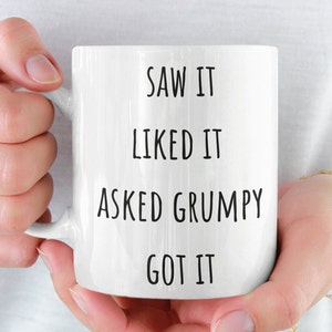 May include: White ceramic mug with black text that reads "SAW IT, LIKED IT, ASKED GRUMPY, GOT IT." The mug is held by two hands. The mug is a standard size for coffee or tea.