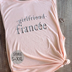 Puede incluir: Un top de tirantes rosa sin mangas con el texto "girlfriend" tachado y "fiancée" escrito debajo. El texto está en gris. El top está disponible en tallas S-XXL.