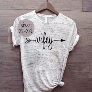 Puede incluir: Una camiseta gris y blanca con una flecha negra y la palabra "wifey" impresa en ella. La camiseta est&aacute; colgada en una percha de madera. La camiseta est&aacute; disponible en tallas XS-XXL.