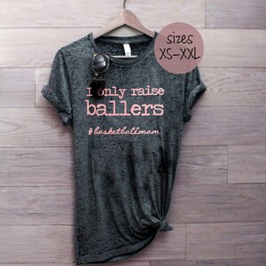 May include: A dark gray t-shirt with the text "I only raise ballers #basketballmom" printed in pink. The shirt is hanging on a wooden hanger against a woodgrain background. Sizes XS-XXL are available.