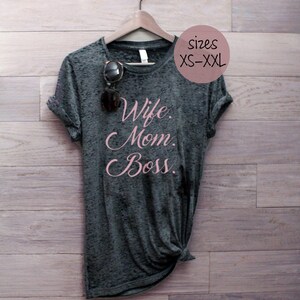 Puede incluir: Camiseta gris con el texto "Wife. Mom. Boss." en letras rosas. La camiseta está colgada de una percha de madera sobre un fondo de madera. Tallas XS-XXL.