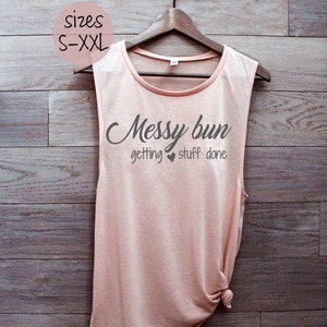 May include: A peach-colored sleeveless tank top with the text "Messy bun getting stuff done" in gray lettering. The tank top is hanging on a wooden hanger against a woodgrain background. The text "sizes S-XXL" is visible in a circle above the tank top.