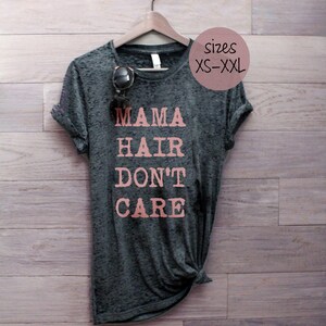 May include: A dark gray t-shirt with the text "MAMA HAIR DON'T CARE" printed in pink. The shirt is hanging on a wooden hanger against a woodgrain background. The text "sizes XS-XXL" is written in pink on a white background.