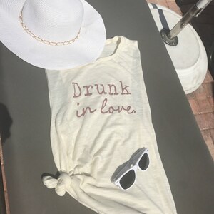 May include: A white tank top with the text "Drunk in love."  The tank top is knotted at the bottom and has a pair of white sunglasses on top of it. A white sun hat is in the background.