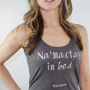 Puede incluir: Un top gris sin mangas con el texto "Na'maste in bed" y el hashtag "#savasana" impreso en blanco.