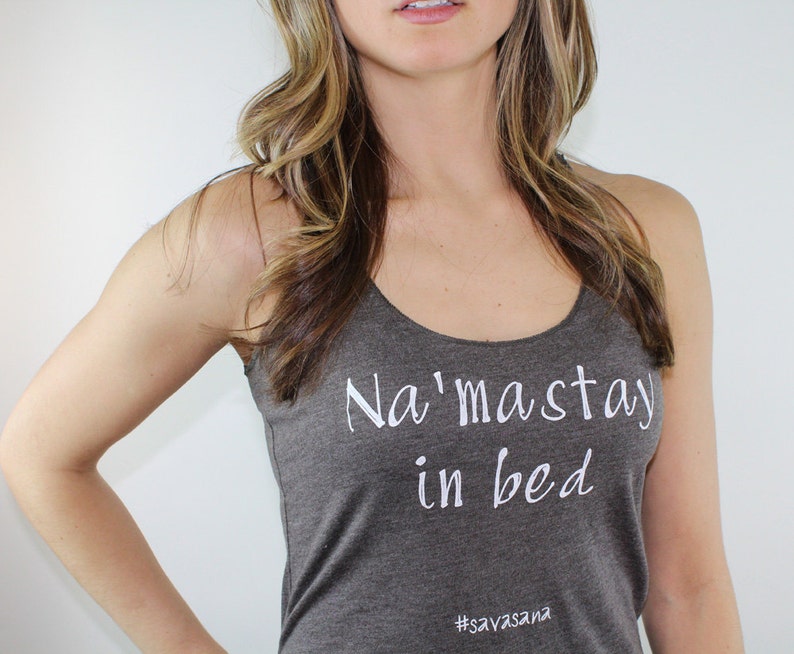 Puede incluir: Un top gris sin mangas con el texto "Na'maste in bed" y el hashtag "#savasana" impreso en blanco.