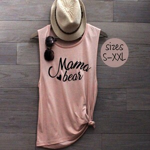 May include: A pink sleeveless tank top with the text "Mama bear" in black lettering and a small heart graphic. The tank top is displayed on a wooden background with a straw hat and sunglasses.