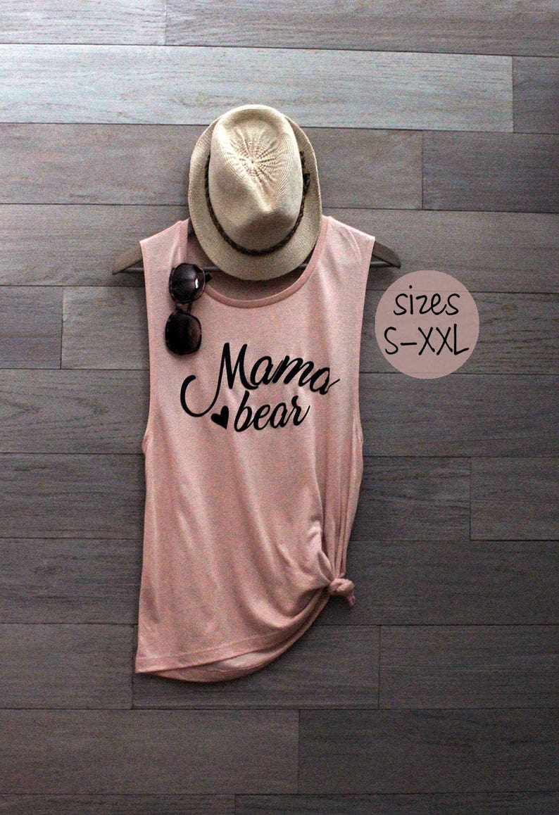 May include: A pink sleeveless tank top with the text "Mama bear" in black lettering and a small heart graphic. The tank top is displayed on a wooden background with a straw hat and sunglasses.
