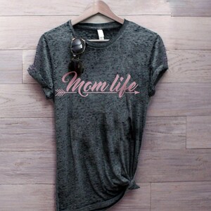 May include: A dark gray t-shirt with a distressed look, featuring the text "Mom Life" in pink script with an arrow pointing to the right.