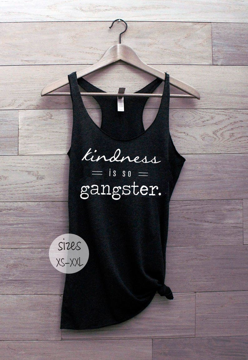 以下が含まれることがあります： 白い文字で「kindness is so gangster.」と書かれた黒いレーサーバックタンクトップ。タンクトップは木製の背景にハンガーでかけられています。サイズXS-XXL。