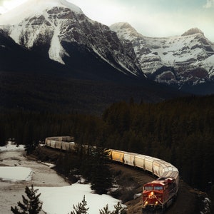 May include: A red freight train with a long line of boxcars winds through a snowy mountain pass. The train is traveling through a forest of evergreen trees. The mountains are covered in snow and the sky is cloudy.