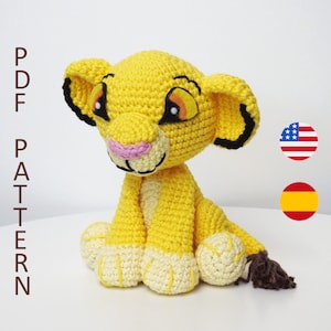 May include: A crocheted stuffed lion cub toy with a yellow body, white paws, and a brown tail. The lion cub has black eyes, a pink nose, and a black outline around its ears.