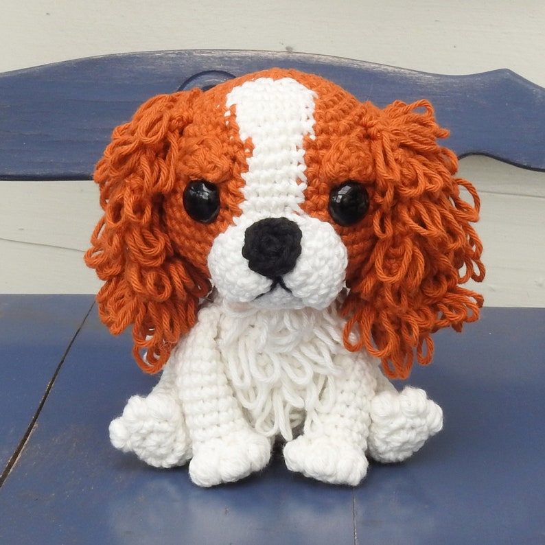 May include: A handmade crochet Cavalier King Charles Spaniel dog. The plush toy features a white and brown head with black eyes and a black nose. The body is white, and the dog is sitting.