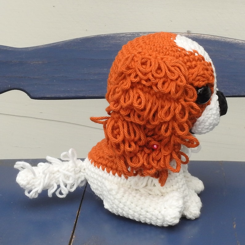 May include: A handmade crochet Cavalier King Charles Spaniel dog. The dog is primarily white with orange and brown accents. The dog has black eyes and a black nose. The dog is sitting on a blue surface.