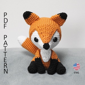 Puede incluir: Un juguete de amigurumi de zorro tejido a crochet con pelaje naranja, pecho blanco y orejas y patas negras. El juguete está sentado sobre una superficie gris. PDF PATTERN ENG