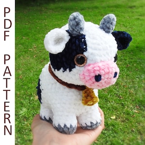 May include: A crocheted plush toy cow with white fur, black spots, and a pink nose. The cow has a brown collar with a gold bell.