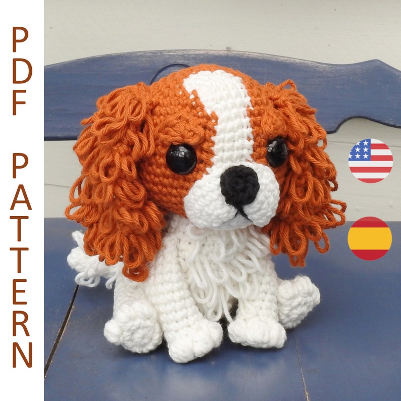 May include: A crocheted Cavalier King Charles Spaniel dog with white, brown, and black details. The dog has large black eyes and a black nose. The text "PDF PATTERN" is on the left side of the image. The dog is sitting on a blue surface.