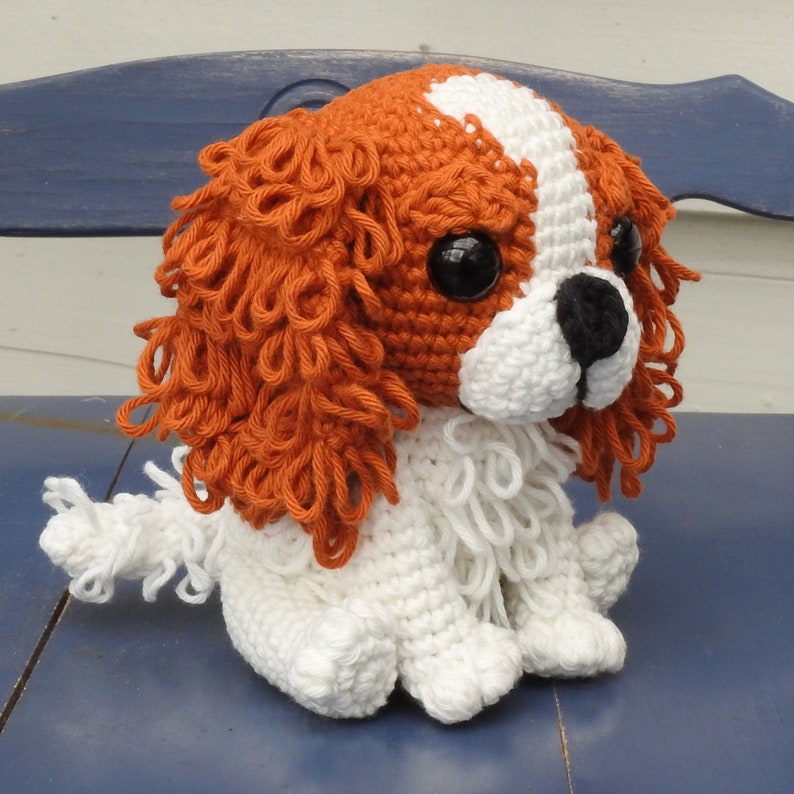 May include: A crocheted Cavalier King Charles Spaniel dog with a white and brown color scheme. The dog has large black eyes, a black nose, and a white stripe on its head. The dog is sitting on a blue surface.