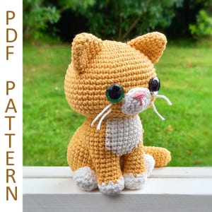 May include: A crocheted orange and white cat with green eyes. The cat is sitting on a white surface.