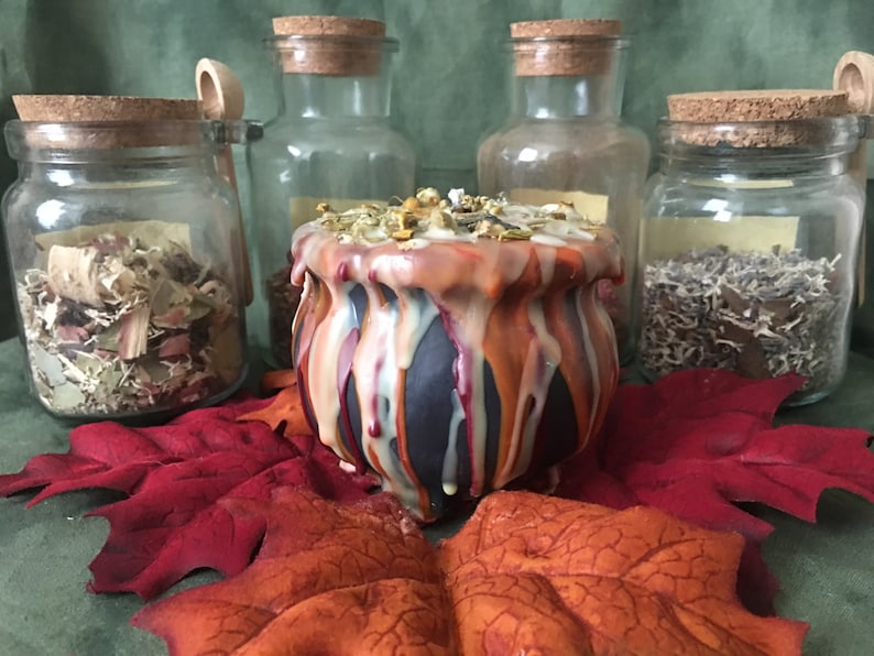 May include: A black cauldron-shaped candle with orange, white, and red wax drips, surrounded by glass jars with cork lids filled with herbs. The candle sits on red and orange artificial leaves, with a green background.