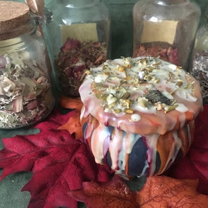 May include: A decorative, round candle with a dripped wax design in orange, white, and blue. The candle is topped with dried herbs and spices. Glass jars filled with dried herbs and flowers are in the background, along with red and orange fall leaves.