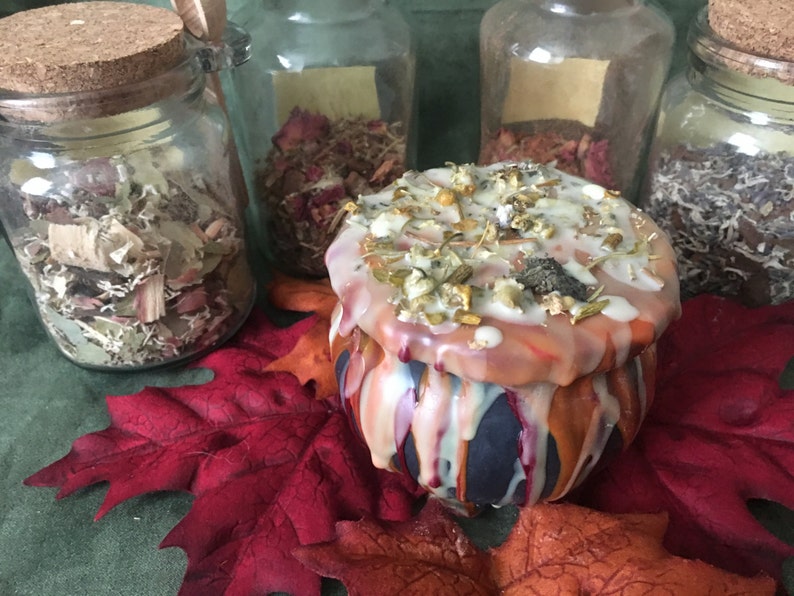 May include: A decorative, round candle with a dripped wax design in orange, white, and blue. The candle is topped with dried herbs and spices. Glass jars filled with dried herbs and flowers are in the background, along with red and orange fall leaves.