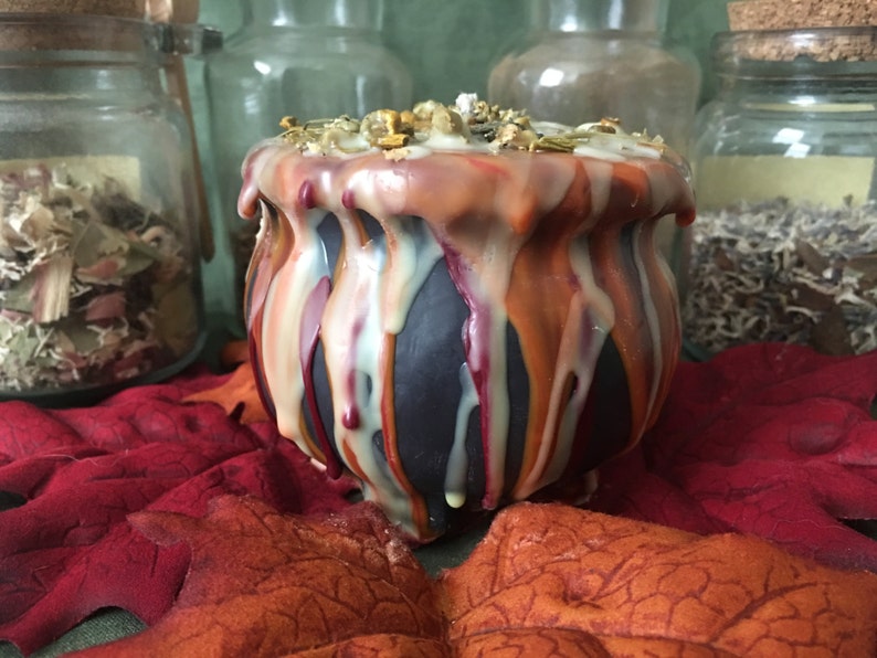 May include: A black cauldron-shaped candle with dripped wax in shades of orange, red, and cream. The candle is decorated with dried herbs and sits on a bed of red and orange leaves, evoking a fall or Halloween theme.