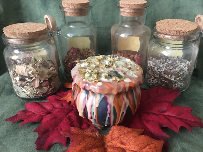 May include: A decorative cauldron-shaped candle with orange, white, and blue wax drips, surrounded by glass jars with cork lids filled with dried herbs and flowers. Red and orange artificial leaves are placed in the foreground.