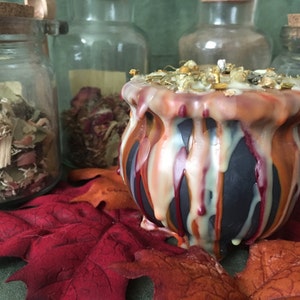 May include: A black cauldron-shaped candle with dripped orange, red, and cream-colored wax. The candle is topped with dried herbs and flowers. The candle sits on a red leaf with glass jars of dried herbs in the background.