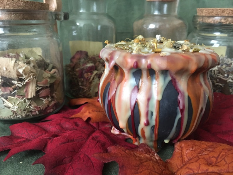 May include: A black cauldron-shaped candle with dripped orange, red, and cream-colored wax. The candle is topped with dried herbs and flowers. The candle sits on a red leaf with glass jars of dried herbs in the background.