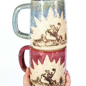 Pode incluir: Duas canecas de cerâmica empilhadas. A caneca superior é azul, a inferior vermelha. Ambas apresentam um desenho de cowboy e cavalo. As canecas têm um acabamento texturizado e uma alça.