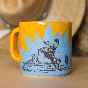May include: A ceramic mug with a speckled yellow top and a light blue base featuring a dark brown graphic illustration of a cowboy riding a bucking horse in a desert landscape with cacti and rock formations under a jagged sunburst design.
