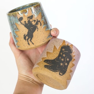 Puede incluir: Dos tazas de cerámica con temática de vaquero. Una taza presenta una silueta negra de un vaquero sobre un caballo encabritado, la otra una bota de vaquero con estrellas. Las tazas tienen una base moteada de color canela con un borde azul y marrón.