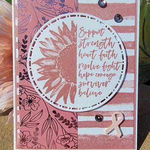 May include: A handmade greeting card with a pink and white color scheme. The card features a sunflower design and the words "Support, strength, heart, faith, resolve, fight, hope, courage, survivor, believe." A pink ribbon is also included.