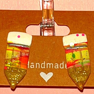 May include: A pair of handmade pencil earrings with a gold glitter finish. The pencils are multi-coloured with pink, yellow, green, and orange stripes. The earrings are displayed on a brown card with the word "handmade" printed on it.