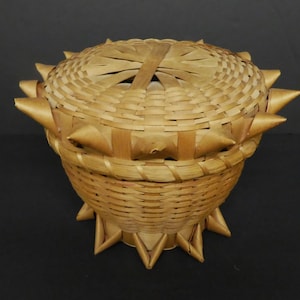 May include: A woven basket with a lid, both decorated with pointed, triangular shapes. The basket is made of natural brown material and has a circular base.