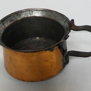 Peut inclure: Une casserole en cuivre avec une poignée en fer noir. La casserole a un fond arrondi et une ouverture large et peu profonde. La poignée est fixée sur le côté de la casserole et se courbe vers le haut et au-dessus du dessus.