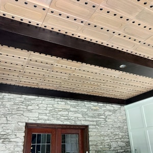 Op de afbeelding: Een plafond met lichtgekleurde rechthoekige houten panelen in een rasterpatroon, geaccentueerd door donkerbruine houten balken. De panelen zijn bevestigd met rijen kleine zwarte ronde bevestigingsmiddelen, wat zorgt voor een geometrisch ontwerp.