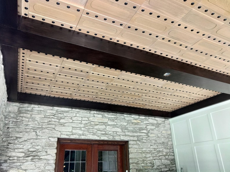 Op de afbeelding: Een plafond met lichtgekleurde rechthoekige houten panelen in een rasterpatroon, geaccentueerd door donkerbruine houten balken. De panelen zijn bevestigd met rijen kleine zwarte ronde bevestigingsmiddelen, wat zorgt voor een geometrisch ontwerp.