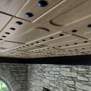 Op de afbeelding: Een plafondinstallatie bestaande uit lichtgekleurde houten panelen met een herhalend patroon van verzonken rechthoekige secties en kleine, donkere ronde accenten die in lineaire rijen over het gehele oppervlak zijn gerangschikt.