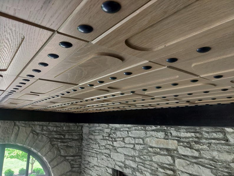 Op de afbeelding: Een plafondinstallatie bestaande uit lichtgekleurde houten panelen met een herhalend patroon van verzonken rechthoekige secties en kleine, donkere ronde accenten die in lineaire rijen over het gehele oppervlak zijn gerangschikt.
