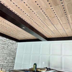Op de afbeelding: Een plafond met lichtgekleurde rechthoekige houten panelen in een rasterpatroon, gescheiden door donkerbruine houten balken. De panelen hebben afgeronde verzonken centra en zijn voorzien van kleine, gelijkmatig verdeelde zwarte bevestigingen.